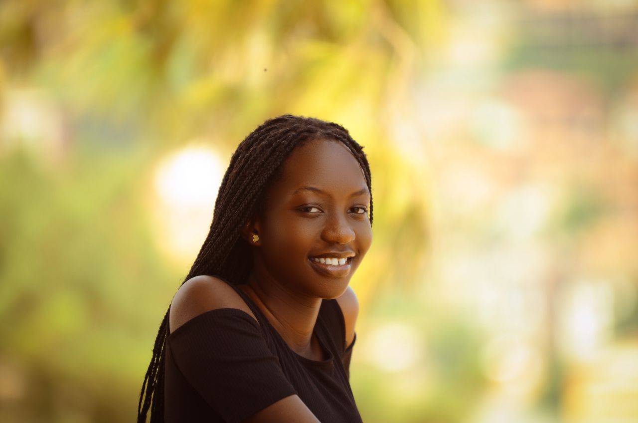 Portrait of a cheerful young woman with braided hair wearing a black top, captured in an outdoor setting with a blurred background.